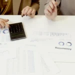 Dua profesional menganalisis dokumen keuangan menggunakan kalkulator di meja kerja